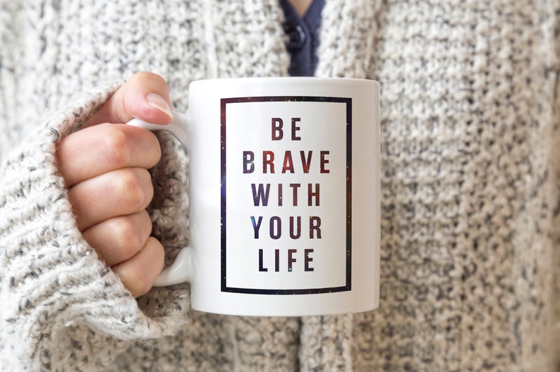 Pode incluir: Caneca de cer&acirc;mica branca com uma borda preta em torno de um fundo semelhante a uma gal&aacute;xia. O texto "BE BRAVE WITH YOUR LIFE" &eacute; impresso em preto.