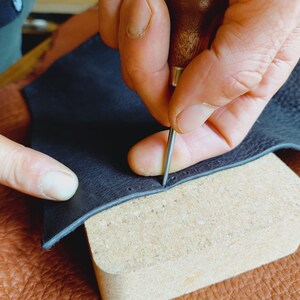 May include: A person using a leather stitching tool to sew a dark blue leather piece. The leather is placed on a light brown cork surface.