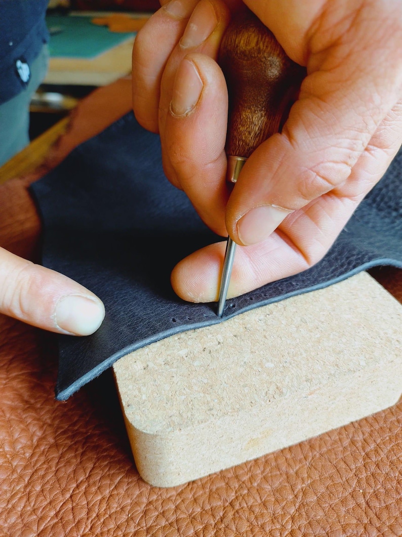 May include: A person using a leather stitching tool to sew a dark blue leather piece. The leather is placed on a light brown cork surface.