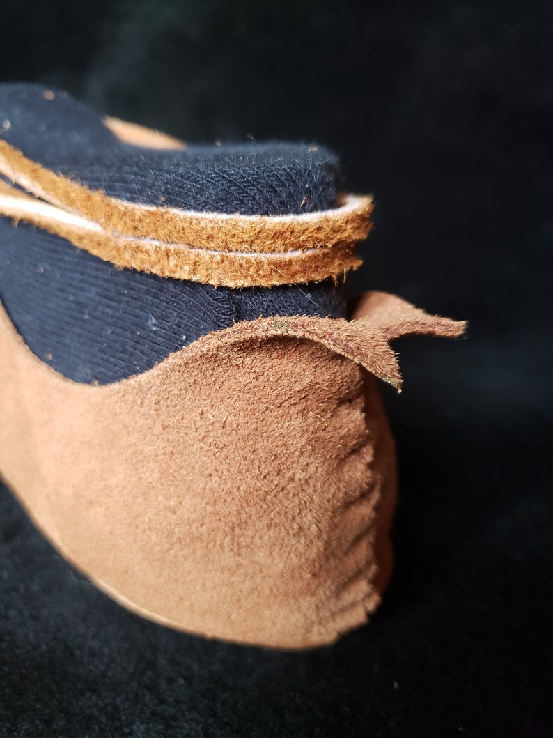 Pode incluir: Close-up de um mocassim de camur&ccedil;a marrom com forro de tecido preto e acabamento em couro marrom. O mocassim est&aacute; dobrado na parte superior, revelando o forro.