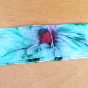 Könnte beinhalten: Ein rechteckiger, handgefärbter Schal mit Aquarelleffekt. Der Schal hat einen hellblauen Hintergrund mit roten und grünen floralen Akzenten. Der Stoff ist der Länge nach gefaltet.