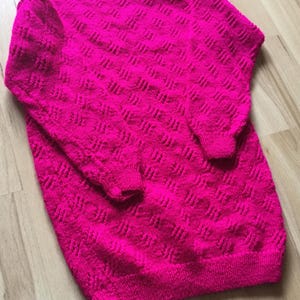 Könnte beinhalten: Ein leuchtend pinkfarbener Strickpullover mit einem strukturierten Rautenmuster. Der Pullover hat lange Ärmel und einen Rundhalsausschnitt.