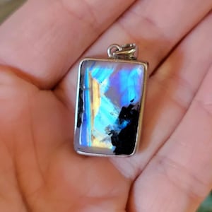 May include: A rectangular pendant featuring a polished gemstone with iridescent blue and gold flashes set in a silver-toned metal frame. The stone displays a smooth surface with deep black inclusions contrasting against the vibrant, glowing light reflections.