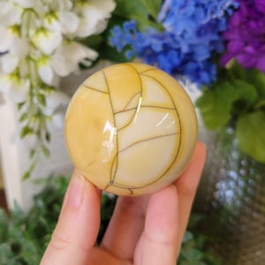 May include: A polished, spherical gemstone with a creamy yellow and tan color palette. The orb features a network of dark, thin lines, creating a unique pattern. The sphere is held in a hand, with a blurred floral background.