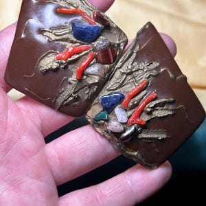 May include: A pair of brown, square-shaped, decorative belt buckles with gold accents and embedded red coral, blue, green, and brown stones.