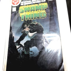 Può includere: Copertina del fumetto Swamp Thing Annual No. 1 con un mostro verde paludoso che tiene una donna con un vestito bianco. La copertina ha il logo DC Comics e il testo "Swamp Thing" in grandi lettere verdi.
