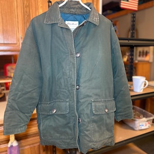 Może przedstawiać: Długa, zielona kurtka z długimi rękawami, zapinana na guziki, z dwoma dużymi kieszeniami. Kurtka ma brązowy kołnierz i niebieską podszewkę. Etykieta głosi "Eddie Bauer".