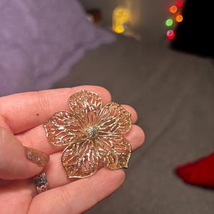 Puede incluir: Un colgante de flor de filigrana dorada con un diseño delicado. El colgante es intrincado y detallado, con una pequeña piedra central.