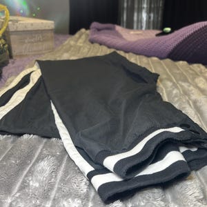Può includere: Pantaloni della tuta neri con strisce bianche sui lati e strisce bianche intorno ai polsini. I pantaloni della tuta hanno una chiusura a bottone.