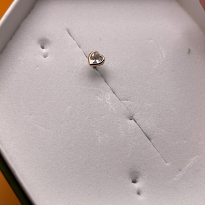 Könnte beinhalten: Ein goldfarbener Ohrstecker mit einem herzförmigen, klaren Edelstein. Der Ohrring ist in Goldmetall gefasst und wird in einer weißen Schachtel präsentiert. Die Schachtel hat eine sechseckige Form und ist mit einem weichen Material ausgekleidet.