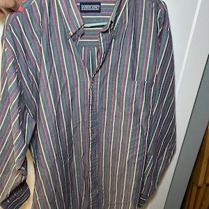 Può includere: Camicia button-down a righe verticali grigie, rosse e gialle. La camicia ha un colletto button-down e una tasca sul petto. Il nome del marchio "LANDS' END" è visibile sull'etichetta.