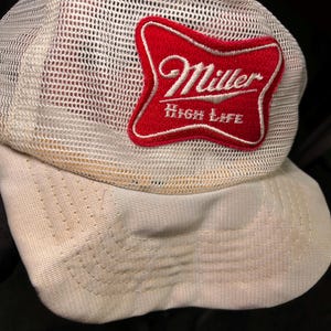 Può includere: Un cappellino da camionista bianco in rete con visiera curva. Una toppa rossa e bianca con il logo Miller High Life è cucita sul davanti. Il cappellino è uno stile classico, adatto per abbigliamento casual.