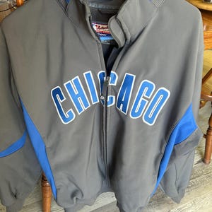 May include: Gray zip-up jacket with blue accents and the word "CHICAGO" in large, white and blue lettering across the chest. The jacket has a stand-up collar and ribbed cuffs. The jacket is hanging on a blue hanger.