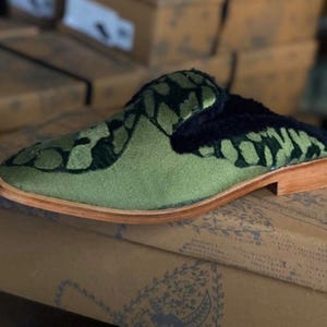 May include: Green velvet slippers with a black and green patterned design. The slippers have a brown leather sole and a black fur lining.