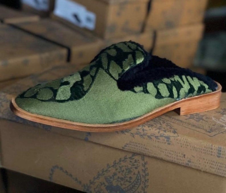 May include: Green velvet slippers with a black and green patterned design. The slippers have a brown leather sole and a black fur lining.