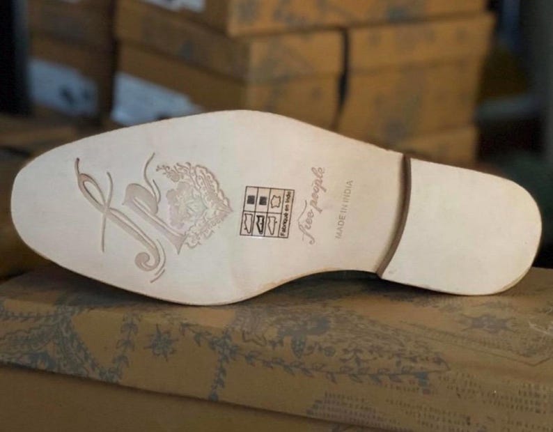 May include: The bottom of a light brown leather shoe with a white sole. The sole has a floral design and the text "Free People" and "Made in India".