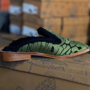 May include: Green velvet slippers with a black fur trim and a wooden sole. The slippers have a pointed toe and a unique patterned design.