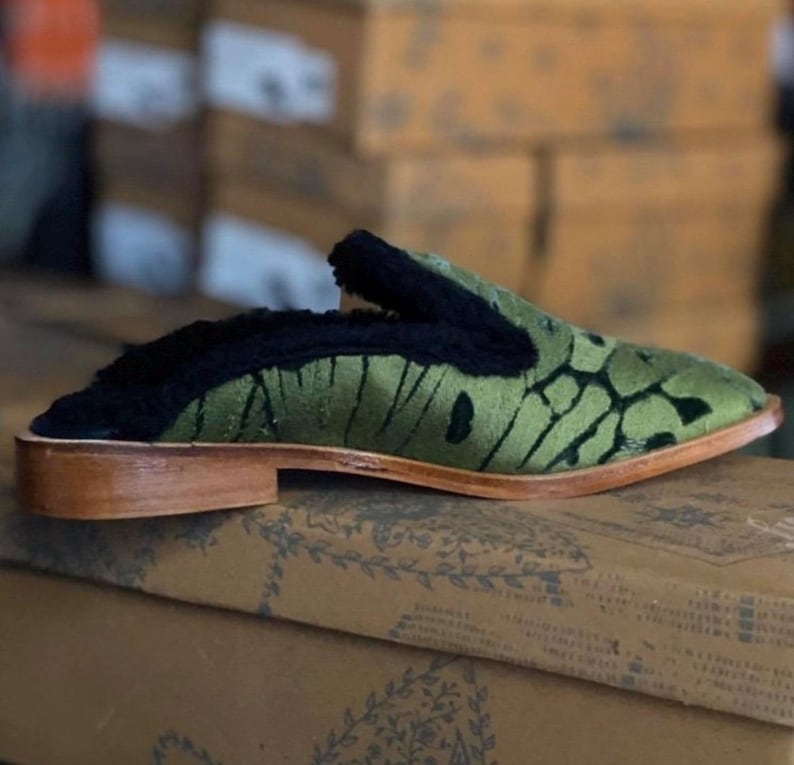 May include: Green velvet slippers with a black fur trim and a wooden sole. The slippers have a pointed toe and a unique patterned design.