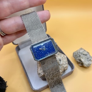 May include: Vintage rectangular watch with a mesh metal strap. The watch face is a deep blue with a speckled pattern. The watch is in a grey box with a light yellow background.