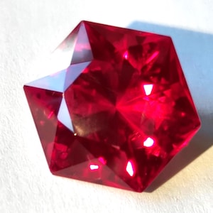Può includere: Una gemma rosso vivo e profondo tagliata in una distinta forma pentagonale con molteplici sfaccettature triangolari. La pietra mostra un'elevata chiarezza e brillantezza, riflettendo la luce attraverso le sue superfici levigate.