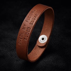 May include: Brown leather bracelet with the embossed text "CHECK MY FUCKING BLOOD SUGAR". The bracelet has a snap closure with a silver-colored button. The background is black.