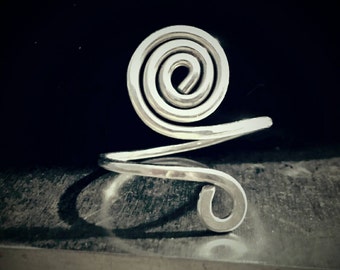 May include: A silver-colored ring with a spiral design. The ring features a coiled spiral at the top, with a band that wraps around the finger. The ring is made of a shiny, metallic material and is set against a dark background.