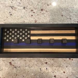 May include: A wooden shadow box with a thin blue line American flag design. The flag is made of wood and has a blue stripe with five black squares.
