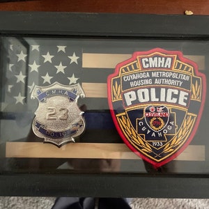 May include: A framed display with an American flag, a blue line, and two badges. The badges are for the Cuyahoga Metropolitan Housing Authority Police. One badge is a silver shield with the number 23 and the words "C.M.H.A. POLICE". The other badge is a red, yellow, and blue patch with the words "CMHA CUYAHOGA METROPOLITAN HOUSING AUTHORITY POLICE" and "CLEVELAND 1796 CUYAHOGA 1933".