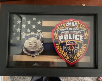 May include: A framed display with an American flag, a blue line, and two badges. The badges are for the Cuyahoga Metropolitan Housing Authority Police. One badge is a silver shield with the number 23 and the words "C.M.H.A. POLICE". The other badge is a red, yellow, and blue patch with the words "CMHA CUYAHOGA METROPOLITAN HOUSING AUTHORITY POLICE" and "CLEVELAND 1796 CUYAHOGA 1933".