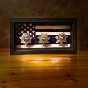 May include: A framed display of three gold and silver police badges with blue ribbons, set against a backdrop of an American flag with a thin blue line.