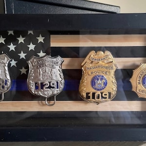 May include: A framed display of four police badges, including two silver badges with the text "City of New York Police" and "Mamaroneck Police", and two gold badges with the text "Mamaroneck Police" and "Sergeant Mamaroneck Police". The badges are displayed on a background of an American flag.