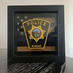 May include: Framed police badge from Kansas, featuring a gold embroidered emblem with the word "POLICE" and "KANSAS". The badge is set against a black and white American flag design with a blue stripe, all encased in a black wooden frame.