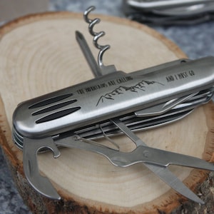 Puede incluir: Una multiherramienta plateada con mango negro y una variedad de herramientas, que incluyen un abridor de botellas, un sacacorchos, un cuchillo y unas tijeras. La herramienta está grabada con el texto "The mountains are calling and I must go".