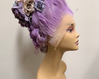 Può includere: Una parrucca viola con una corona di fiori composta da fiori rosa pastello, blu e grigi. La parrucca è acconciata a bouffant con ricci. La parrucca è su una testa di manichino.