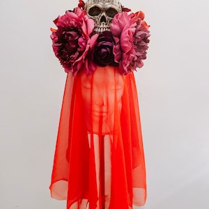 May include: A red sheer veil with a skull and floral headpiece. The skull is white with intricate details and the flowers are pink and purple.