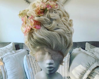 May include: A blonde wig with a large, elaborate updo styled with pink flowers. The wig is on a mannequin head.