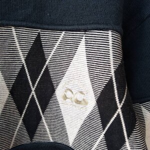 May include: Close-up of a navy blue sweatshirt with a black and white argyle pattern. The argyle pattern is on the lower portion of the sweatshirt. The sweatshirt has a ribbed neckline and a small logo on the argyle pattern.