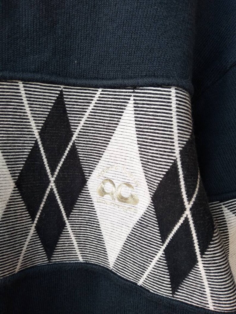 May include: Close-up of a navy blue sweatshirt with a black and white argyle pattern. The argyle pattern is on the lower portion of the sweatshirt. The sweatshirt has a ribbed neckline and a small logo on the argyle pattern.