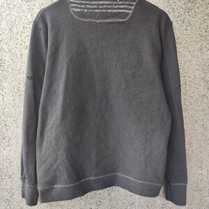 May include: A dark gray, long-sleeved sweatshirt with a ribbed texture. The sweatshirt has a square neckline with three horizontal stripes across the upper back. A small button detail is on the left sleeve. The garment is hanging on a wooden hanger.
