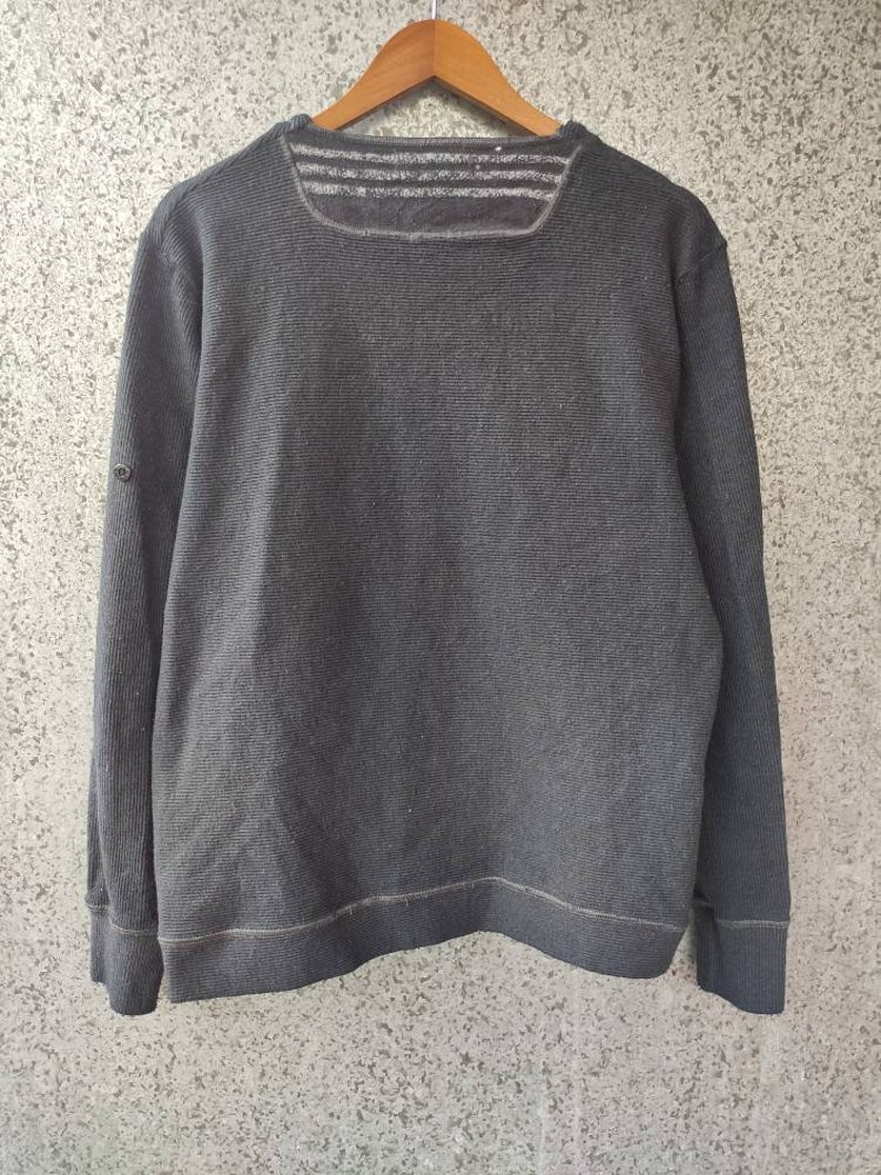 May include: A dark gray, long-sleeved sweatshirt with a ribbed texture. The sweatshirt has a square neckline with three horizontal stripes across the upper back. A small button detail is on the left sleeve. The garment is hanging on a wooden hanger.
