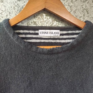 May include: Close-up of a dark gray ribbed knit sweater hanging on a wooden hanger. The collar features a striped pattern in gray and white, with a label that reads "STONE ISLAND".