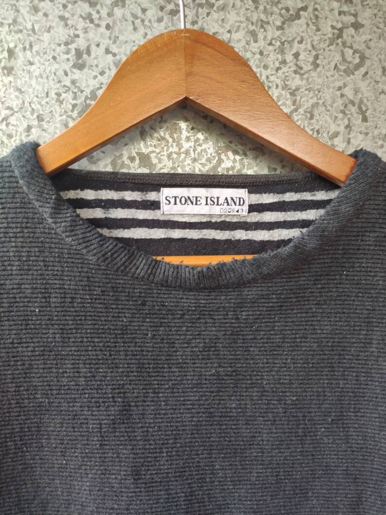 May include: Close-up of a dark gray ribbed knit sweater hanging on a wooden hanger. The collar features a striped pattern in gray and white, with a label that reads "STONE ISLAND".