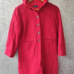 May include: A vibrant red hooded coat with three buttons and two front pockets. The coat is hanging on a wooden hanger. The label reads "agnes b. vintage".