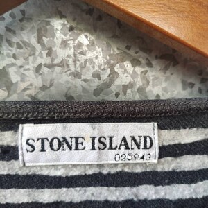 May include: Close-up of a gray and white striped shirt with a Stone Island label. The label is white with black text and the number 025943. The shirt is hanging on a wooden hanger.