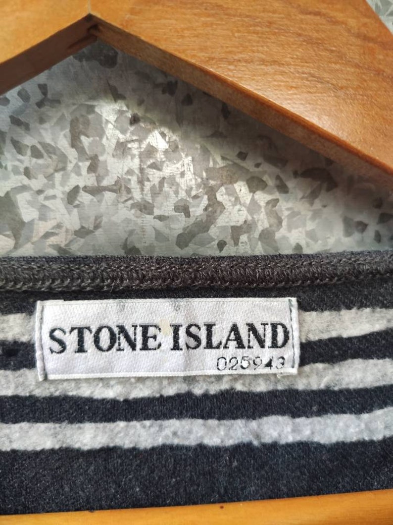 May include: Close-up of a gray and white striped shirt with a Stone Island label. The label is white with black text and the number 025943. The shirt is hanging on a wooden hanger.