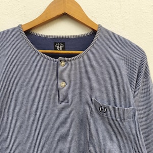 Puede incluir: Camisa de manga larga con estampado azul y blanco con cuello Henley y un bolsillo. La camisa tiene una tapeta abotonada y un cuello azul oscuro. El logotipo de la marca es visible en el cuello y el bolsillo.
