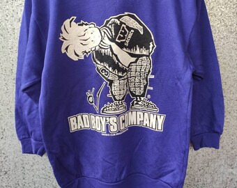 May include: A purple sweatshirt with a white graphic of a cartoon character wearing a baseball cap with the word "Sachico" on it. The graphic also includes the text "Bad Boy's Company" and "Badico Club Production".