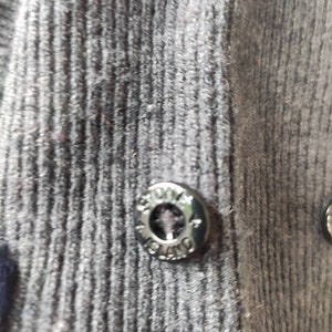 May include: Close-up of a dark gray button with the text "STONE ISLAND" around the edge. The button is attached to a dark gray ribbed fabric. The button is metallic and has a circular design.