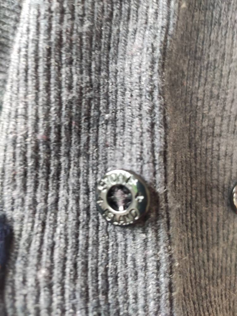 May include: Close-up of a dark gray button with the text "STONE ISLAND" around the edge. The button is attached to a dark gray ribbed fabric. The button is metallic and has a circular design.