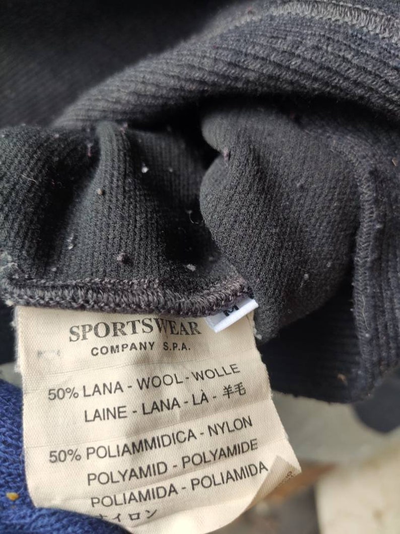 May include: Close-up of a dark gray knit fabric with a ribbed texture. A tag is attached, displaying text in multiple languages, including "SPORTSWEAR COMPANY S.P.A." and fabric composition details: 50% wool and 50% nylon.